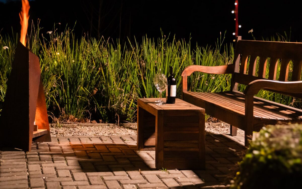 Espaço noturno com deck de madeira, banco de madeira, garrafa e taça de vinho, plantas altas ao fundo, fogueira acesa e luzes vermelhas ao topo