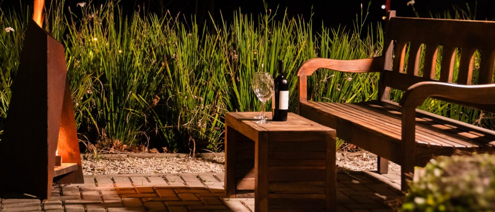 Espaço noturno com deck de madeira, banco de madeira, garrafa e taça de vinho, plantas altas ao fundo, fogueira acesa e luzes vermelhas ao topo
